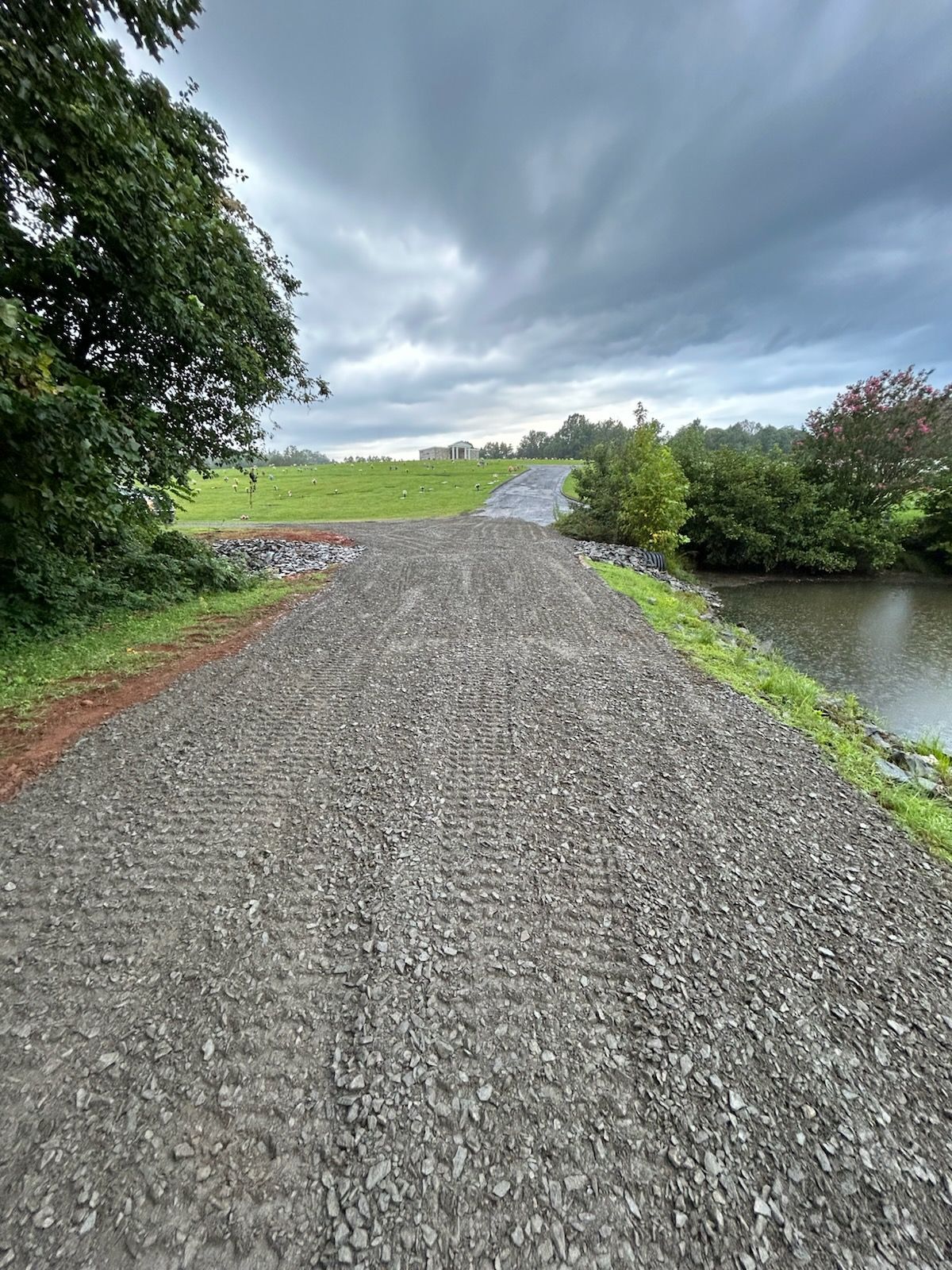 Gravel path beside a pond leading to a grassy field under cloudy skies