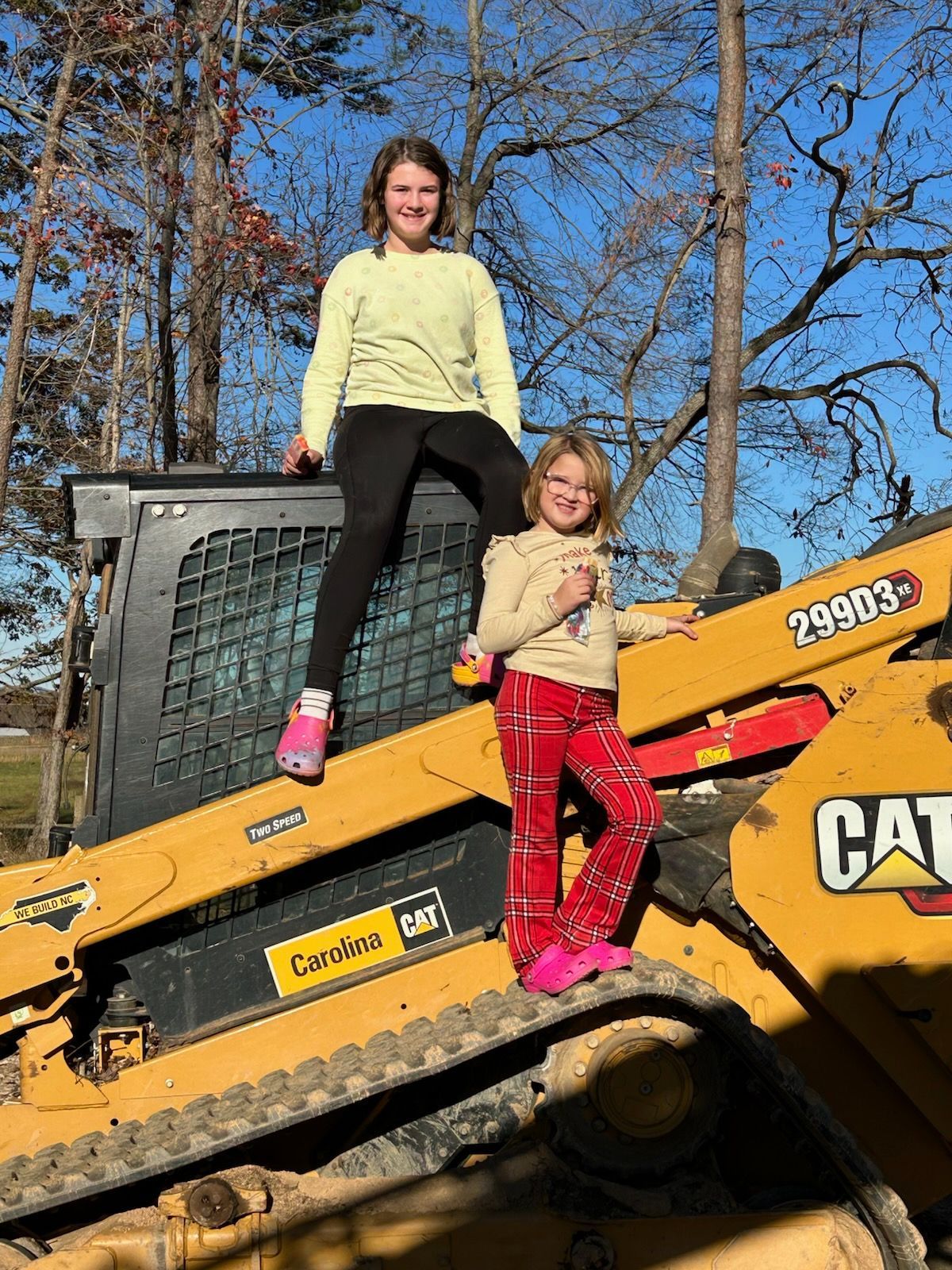 Two children posing on a yellow CAT bulldozer outdoors on a sunny day.