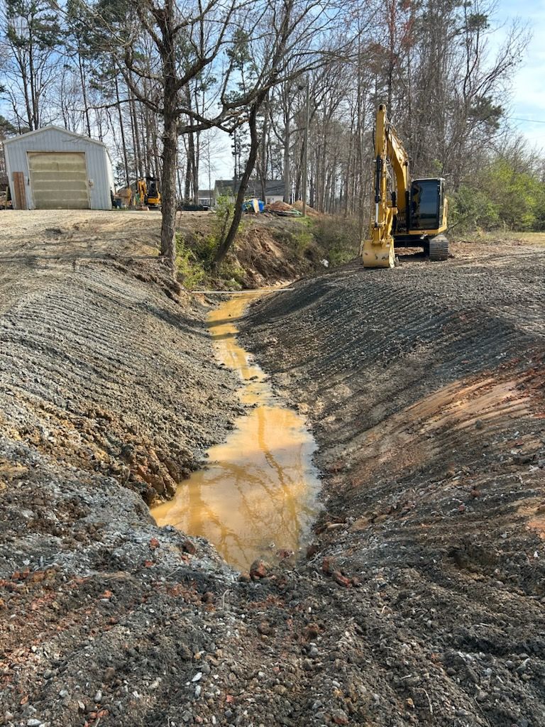 Excavator beside muddy drainage ditch with standing water in a gravel worksite