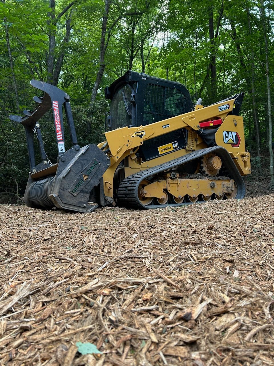 Yellow skid steer with mulcher attachment clearing wood chips in a wooded area