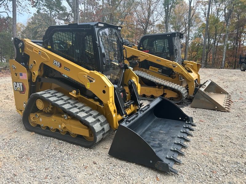 Two yellow CAT tracked skid steer loaders parked on gravel with buckets, autumn trees behind.