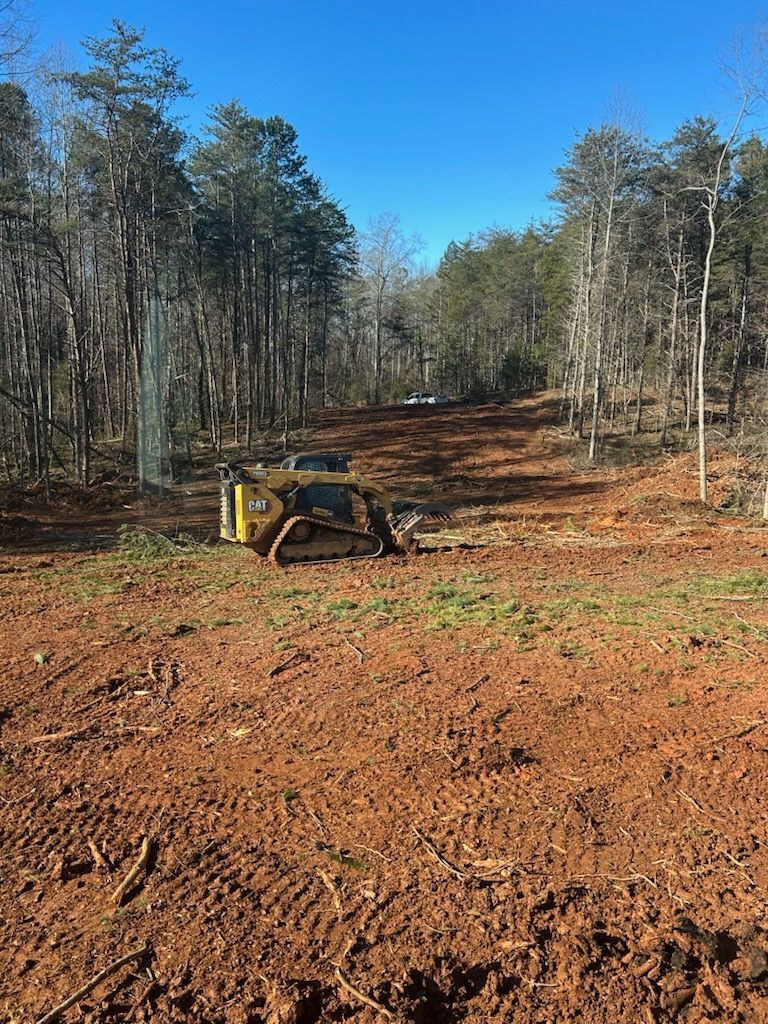 Bulldozer clearing a red dirt path through a wooded area under a clear blue sky