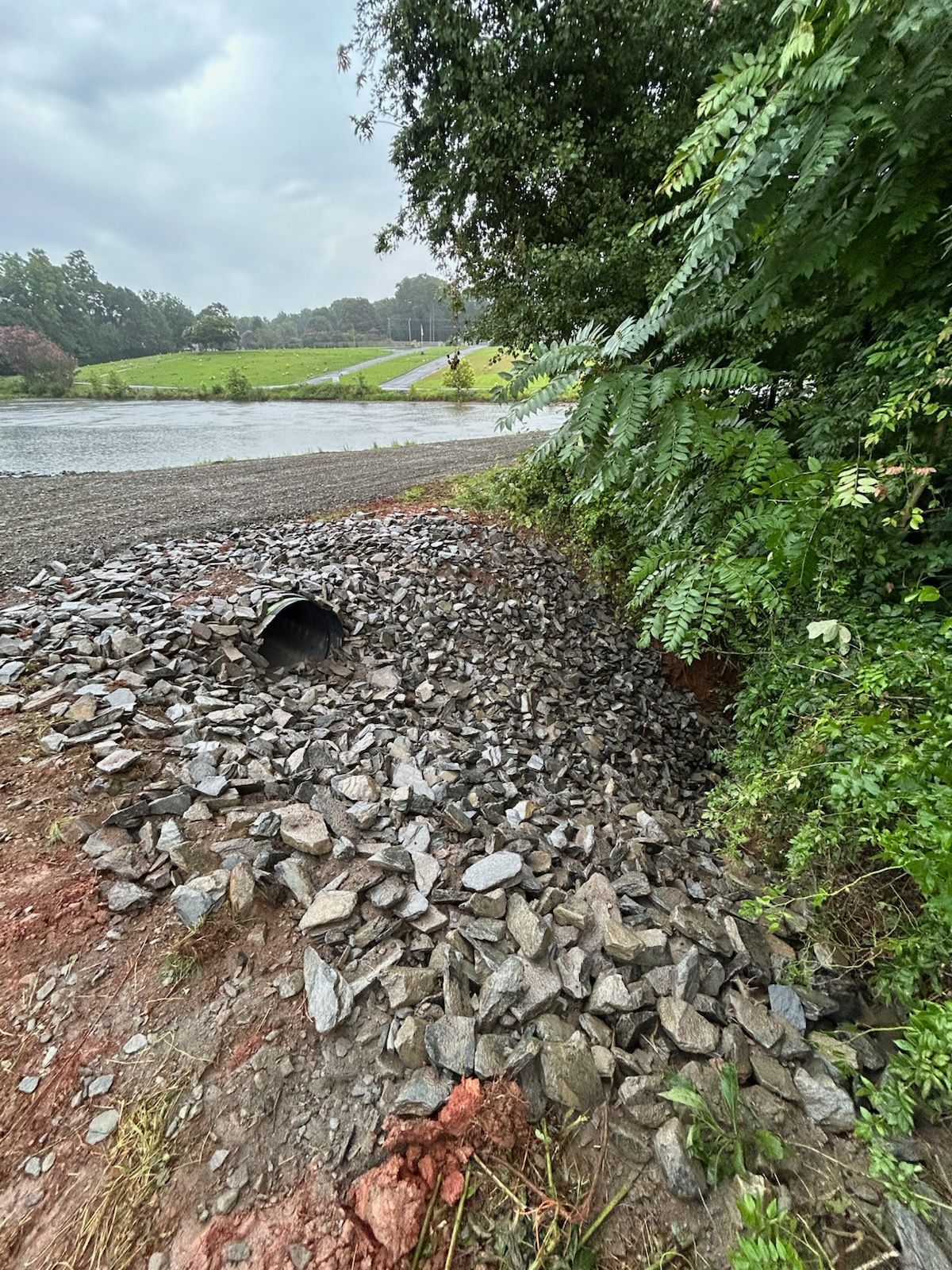Rock pile with a drainage pipe beside a road, with green trees and a pond in the background.