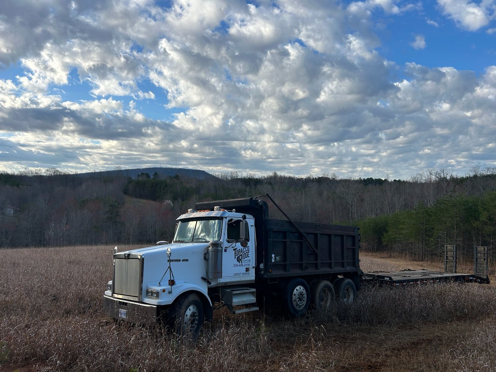 White dump truck in a harvested field under a cloudy sky, with forested hills in the distance