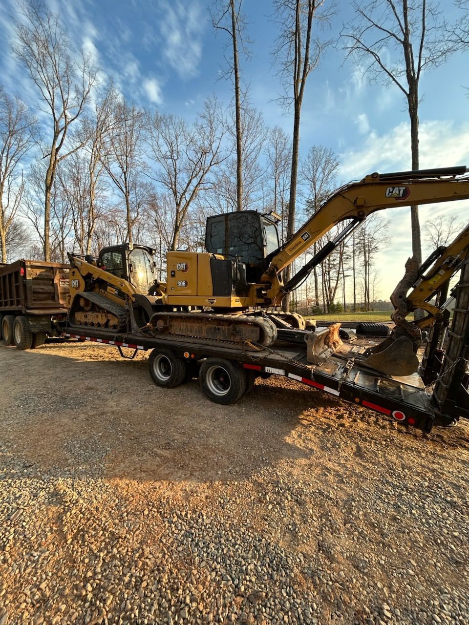 Yellow excavator loaded on a flatbed trailer in a gravel yard with bare trees in the background