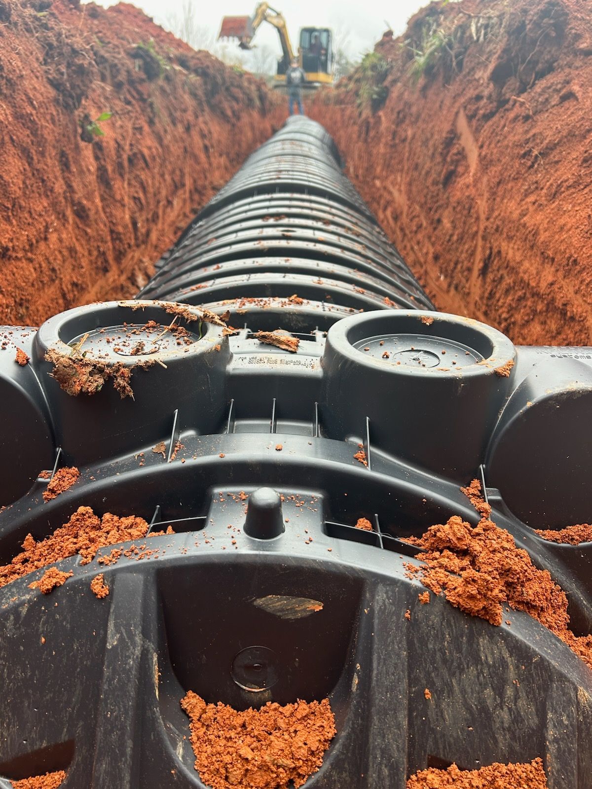 Heavy black pipeline laid in a red dirt trench, with an excavator working in the background.