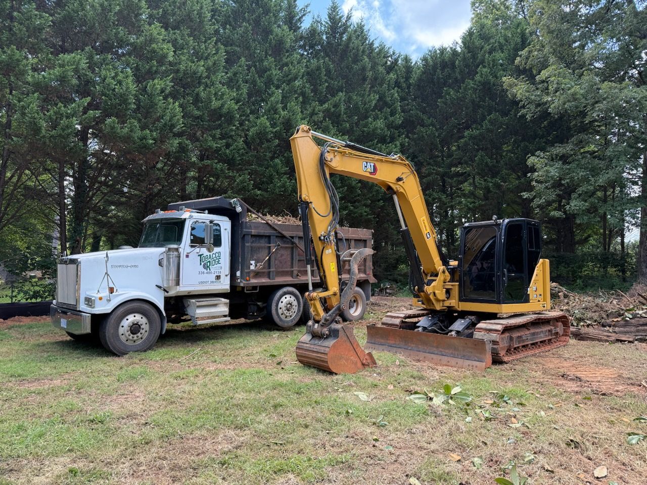 White dump truck and yellow excavator parked on a grassy clearing beside trees