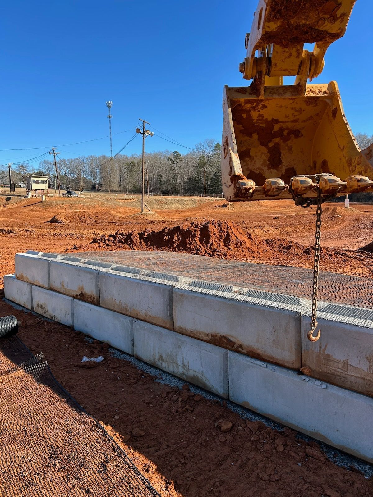 Excavator bucket lowering concrete retaining wall blocks at a red dirt construction site under blue sky