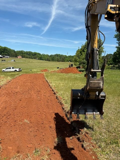 Excavator digging a trench beside a red dirt path in a grassy field under a blue sky