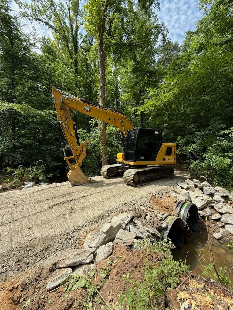 Yellow excavator on a gravel road beside a stone culvert in a wooded area