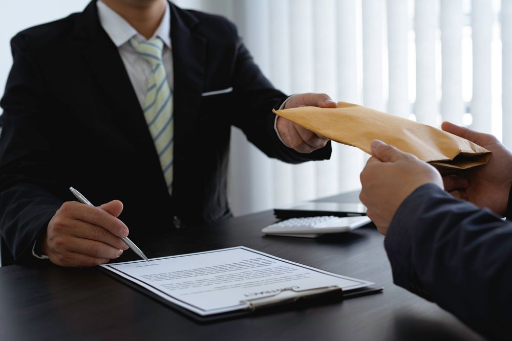 Two men in suits, one handing an envelope to the other while signing a document at a desk.