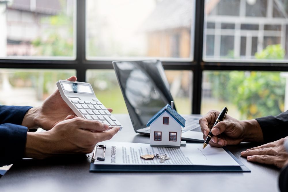 Two people at a table with a house model, calculator, and documents, one signing.