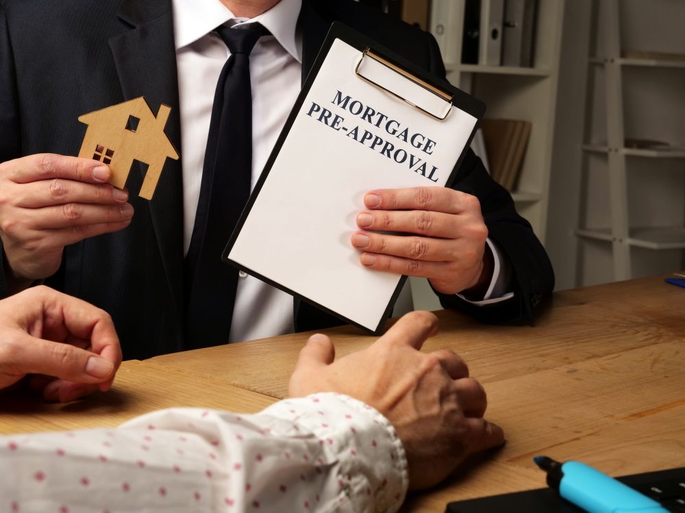 Person in suit holds house cutout and mortgage pre-approval form, presenting to another person at a desk.