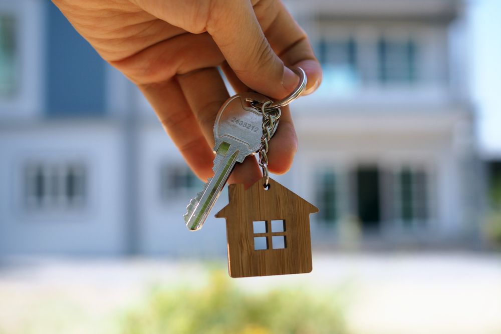 Hand holding house keys with a house-shaped keychain in front of a blurred house.