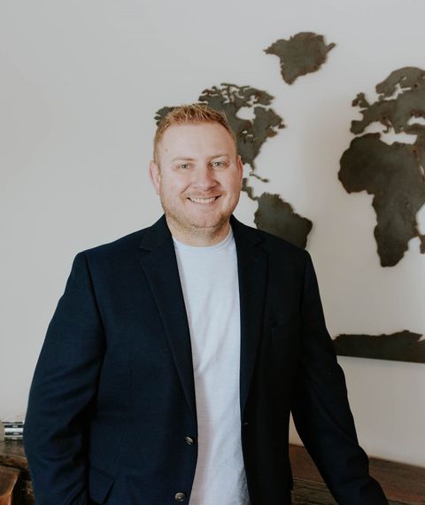 Man in a dark blazer and white shirt standing indoors beside a world map wall decor