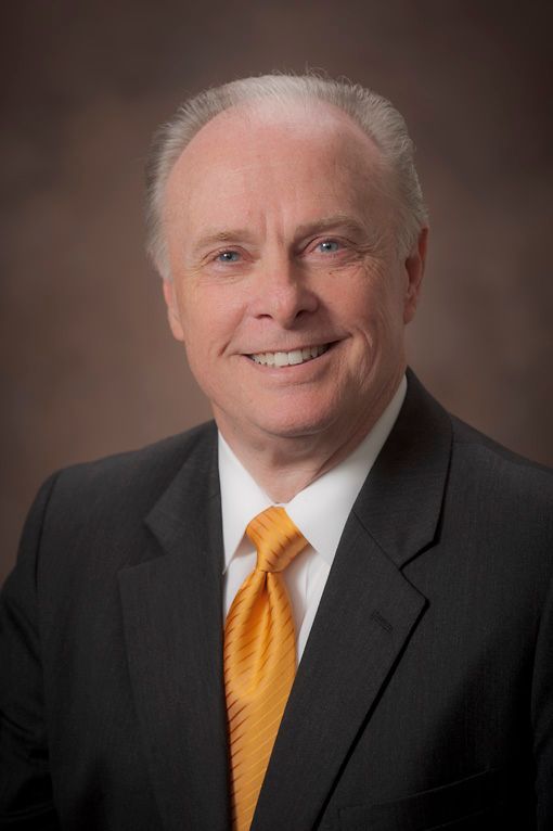 Dr Mark Rutland in suit smiles, orange tie, against a brown gradient background.