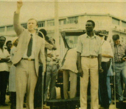 Dr. Mark Rutland in suit speaks to crowd with raised arm; others stand nearby.