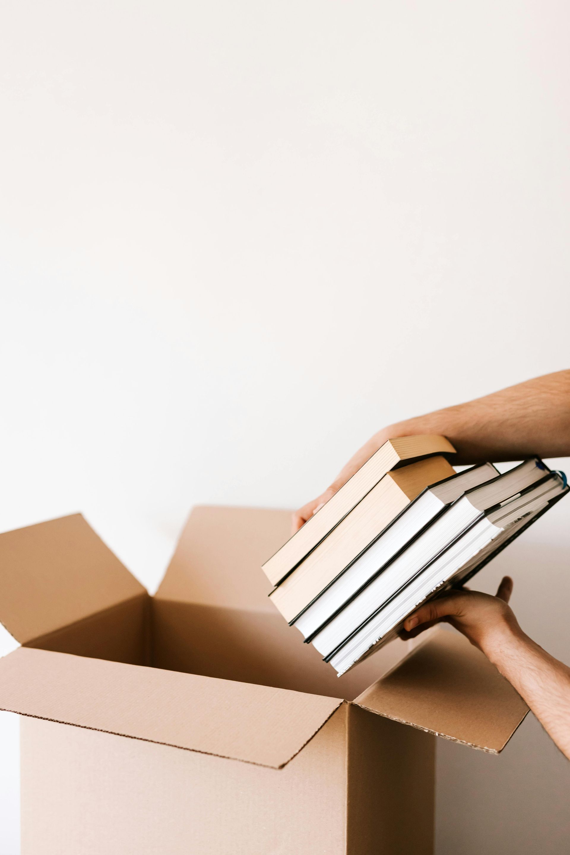 Hands placing stacked books into an open cardboard box on a white background