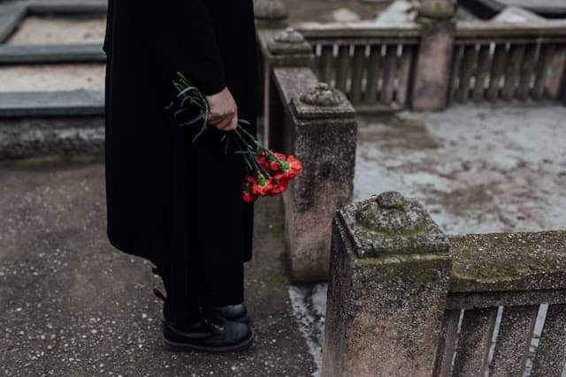A man in a black coat is holding a bouquet of red roses in a cemetery.