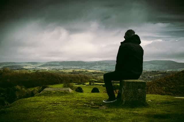 A man is sitting on a stump on top of a hill looking at the landscape.