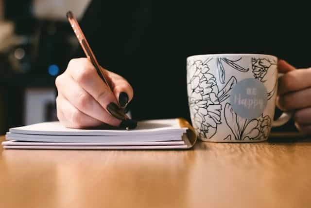 A person is writing in a notebook next to a cup that says be happy.