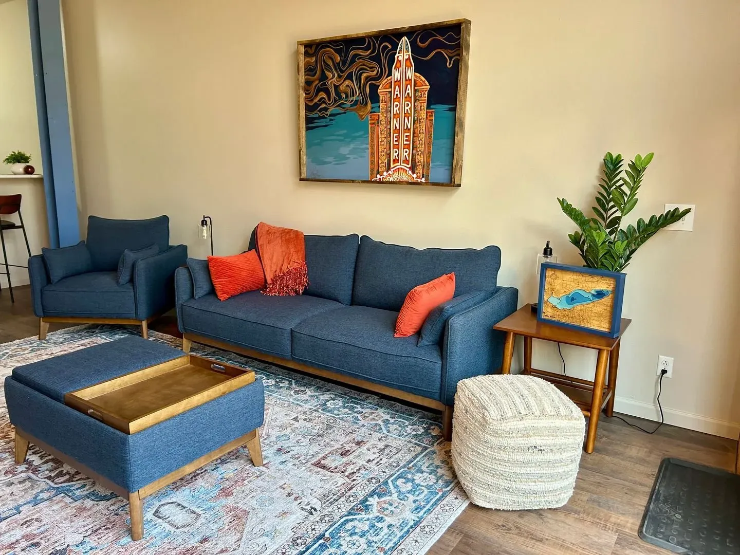 Living room with blue sofa, orange pillows, ottoman, and artwork; rug on the floor.