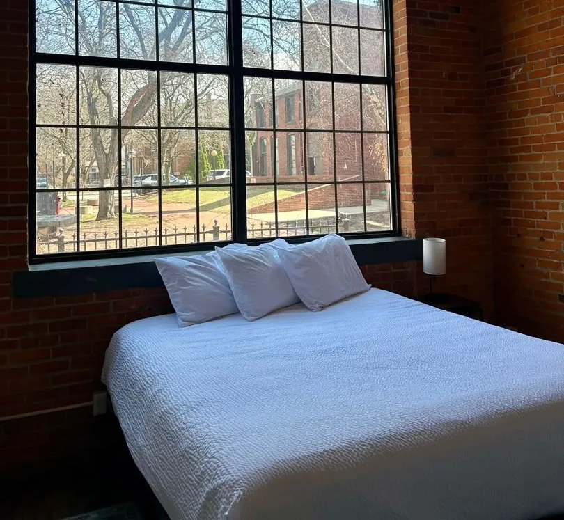 Bed with white bedding and pillows by a large window in a brick-walled room, outside view.