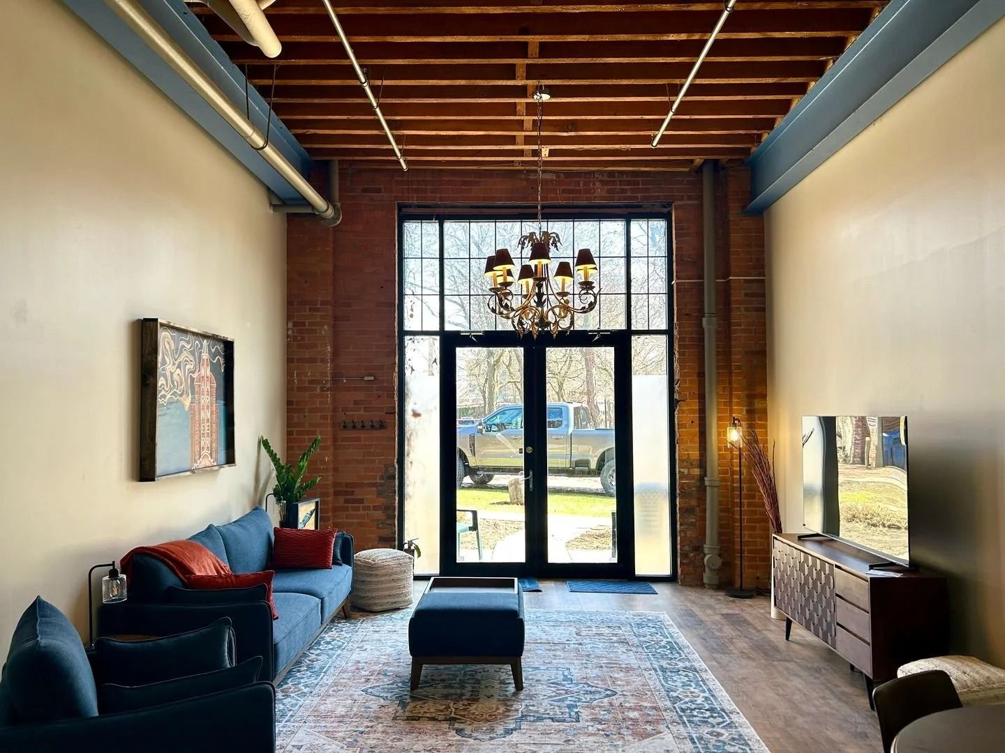 Living room with exposed brick and beams, navy sofa, and chandelier. Large glass doors lead outside.