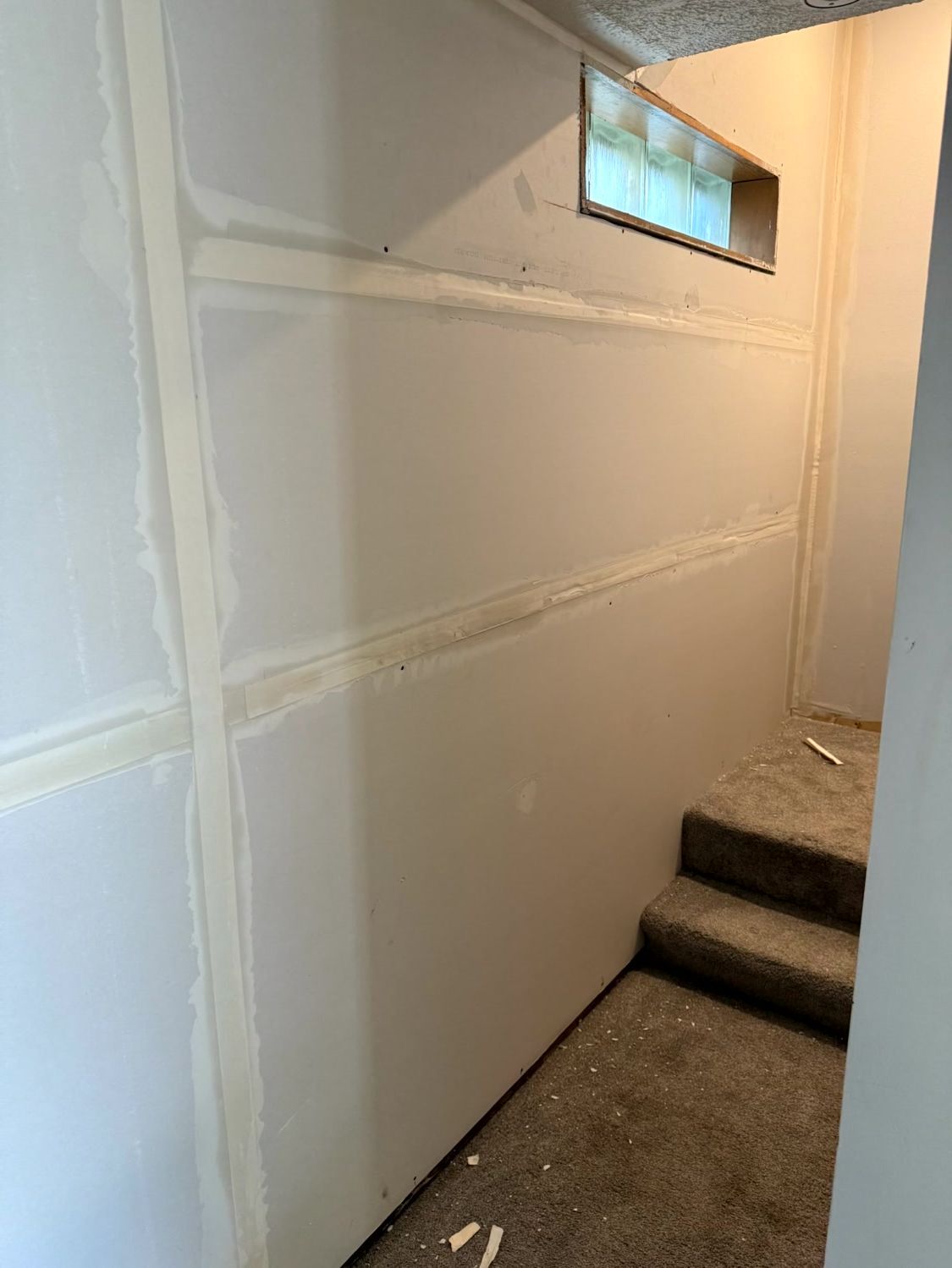 A staircase in a basement with light-colored, unfinished drywall panels that have seams taped with joint compound.