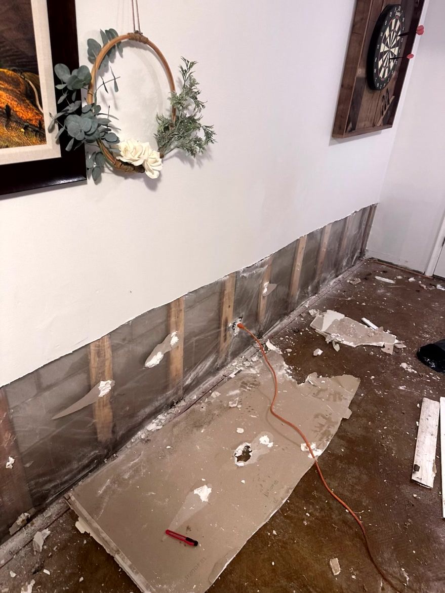 A room with a damaged lower wall where drywall has been removed to reveal wood studs, with debris on the floor.