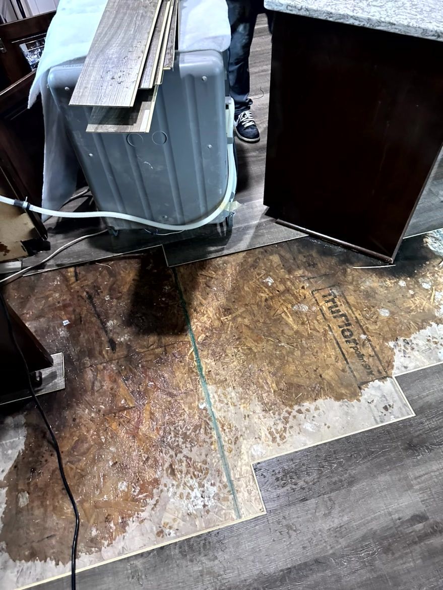 A work area showing an exposed subfloor with dark water damage, planks of flooring, and a portable dehumidifier.