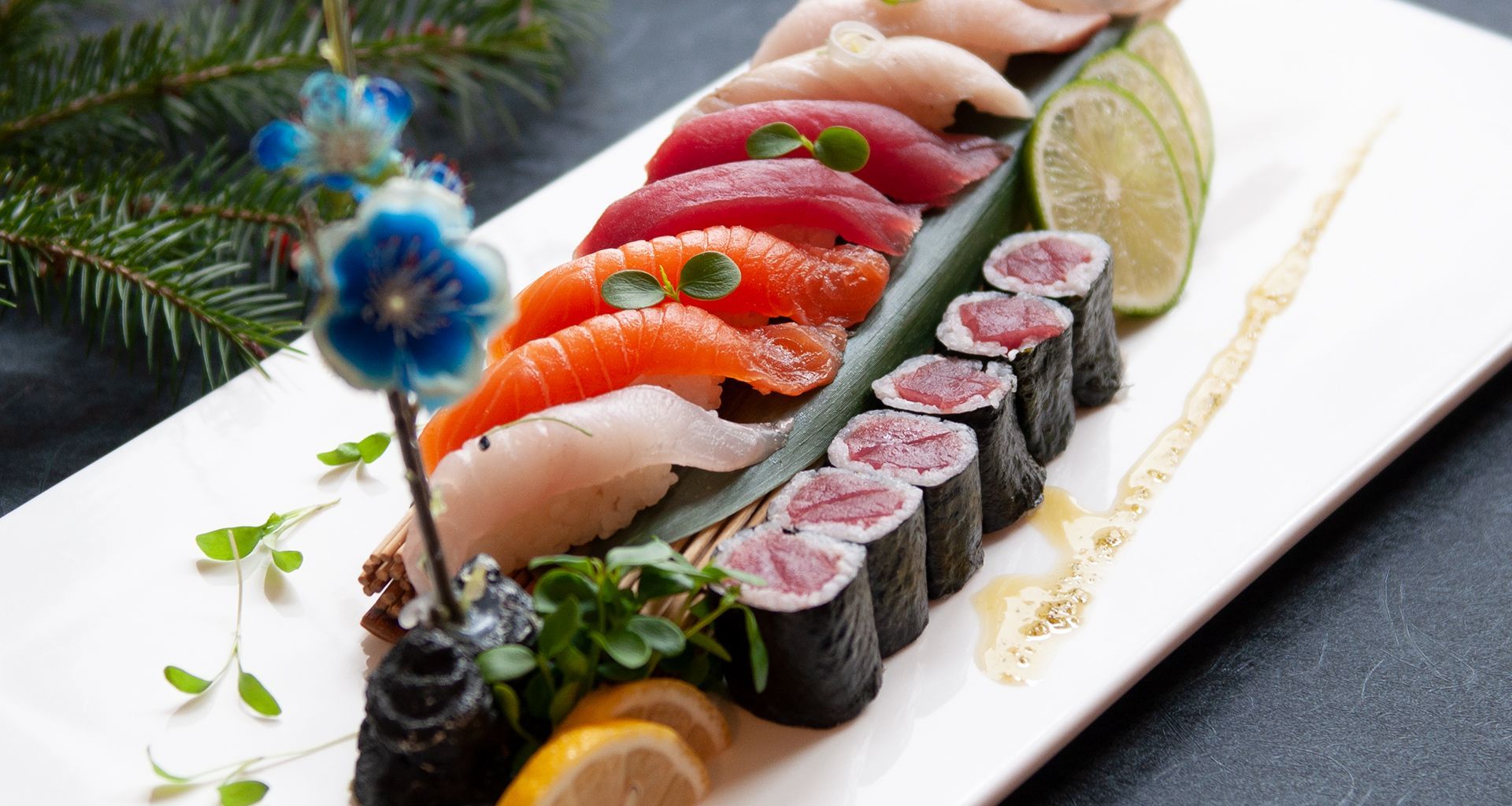 Assorted sushi rolls and nigiri on a white platter, garnished with lime, flowers, and greens.
