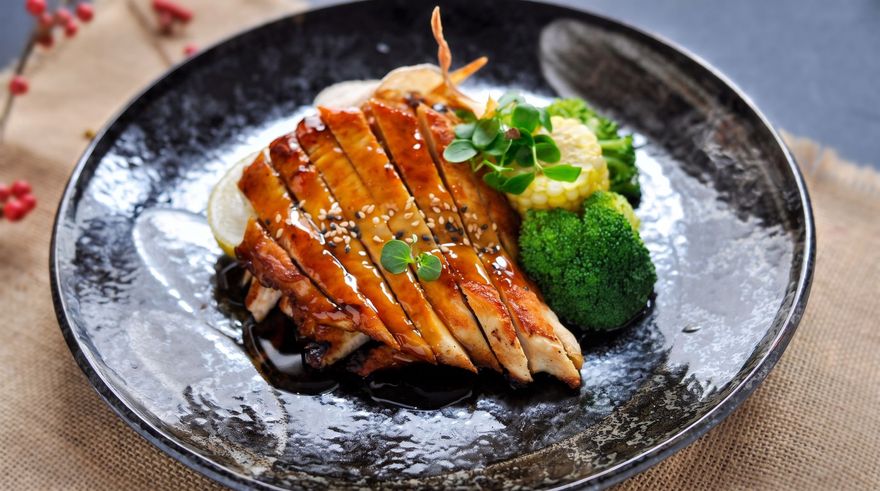 Sliced glazed chicken on a dark plate with broccoli and greens, garnished with sesame seeds