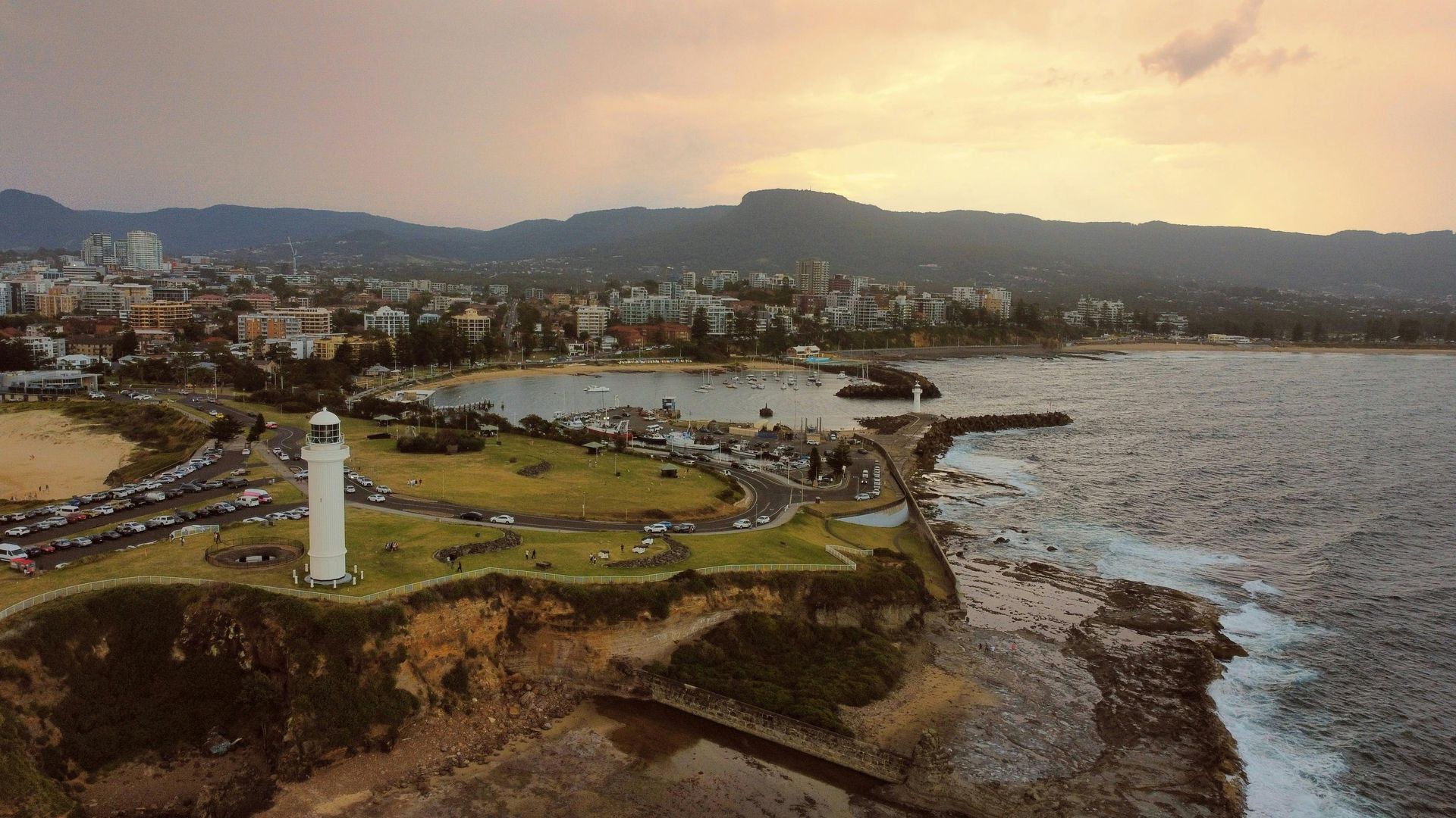 Coastal lighthouse at sunset overlooking rocky shoreline, grassy park, and distant town and mountains