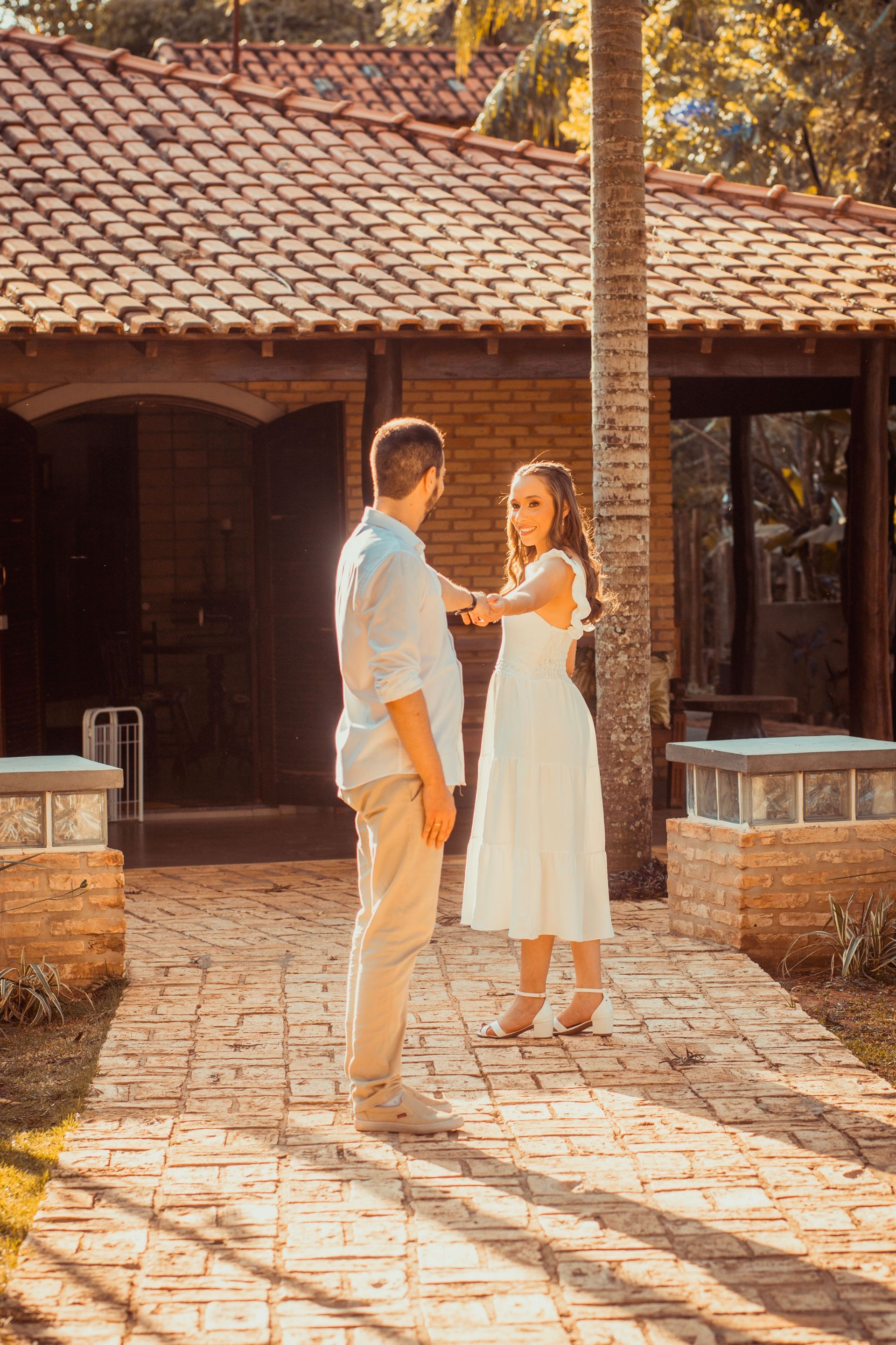 Two people talking in a sunlit courtyard beside a rustic brick building with a tiled roof.