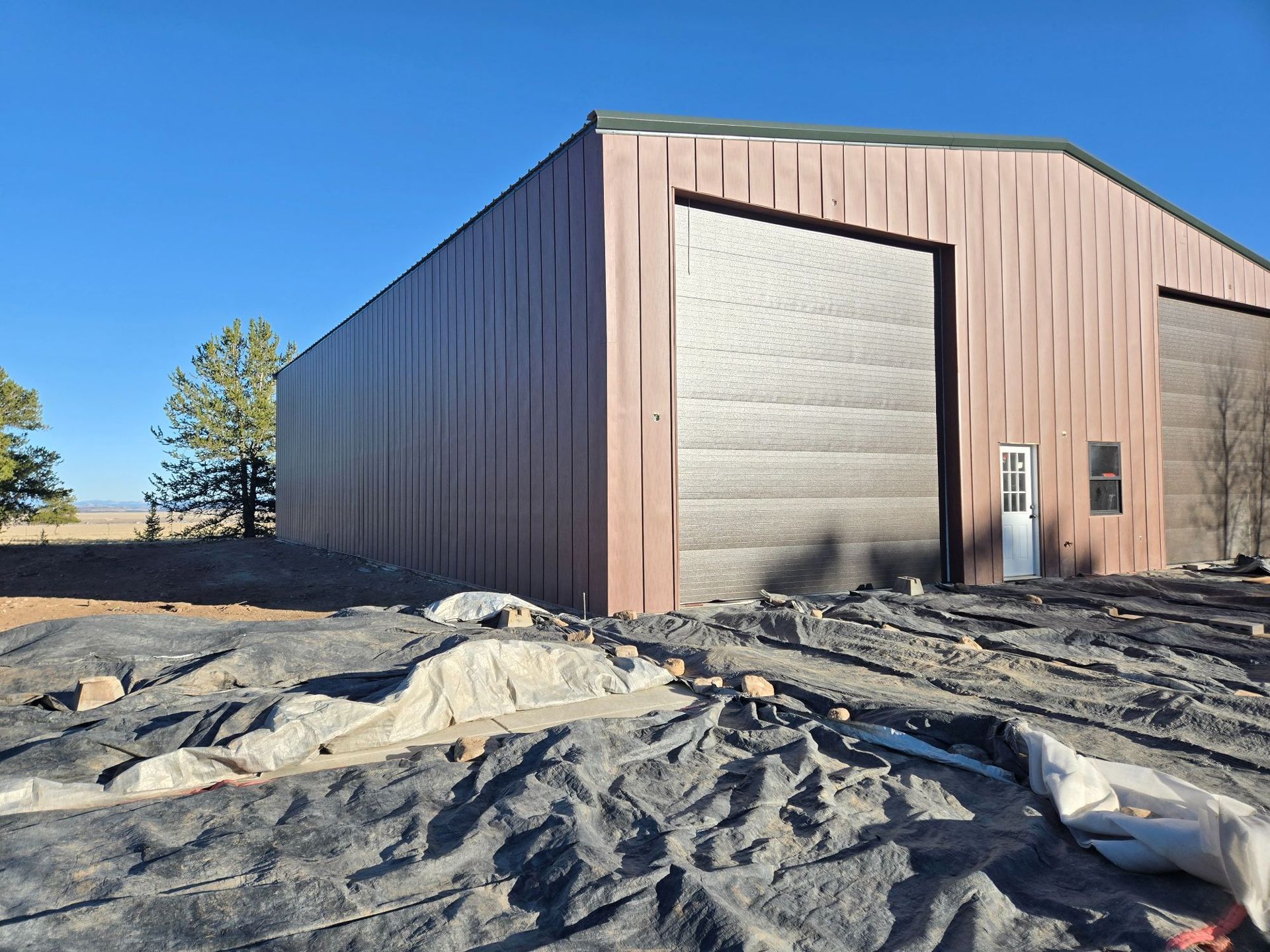 Large metal warehouse with a roll-up door on a dirt lot under a clear blue sky.