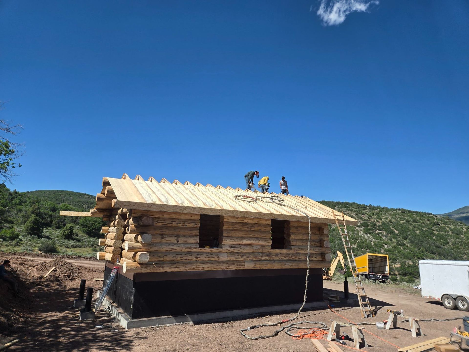 Partially built log cabin under a bright blue sky, with a roof under construction in a rural area