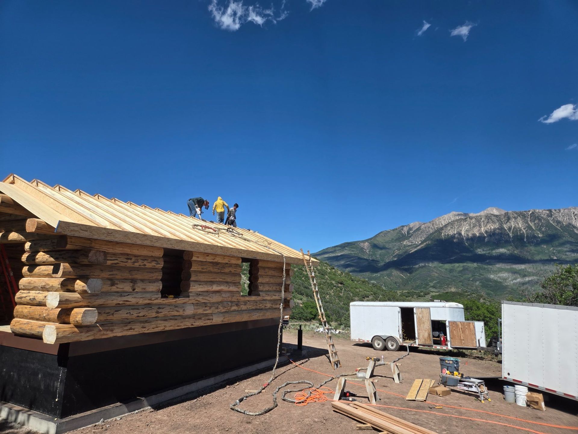 Log cabin under construction with workers on the roof, mountains and trailers in the background