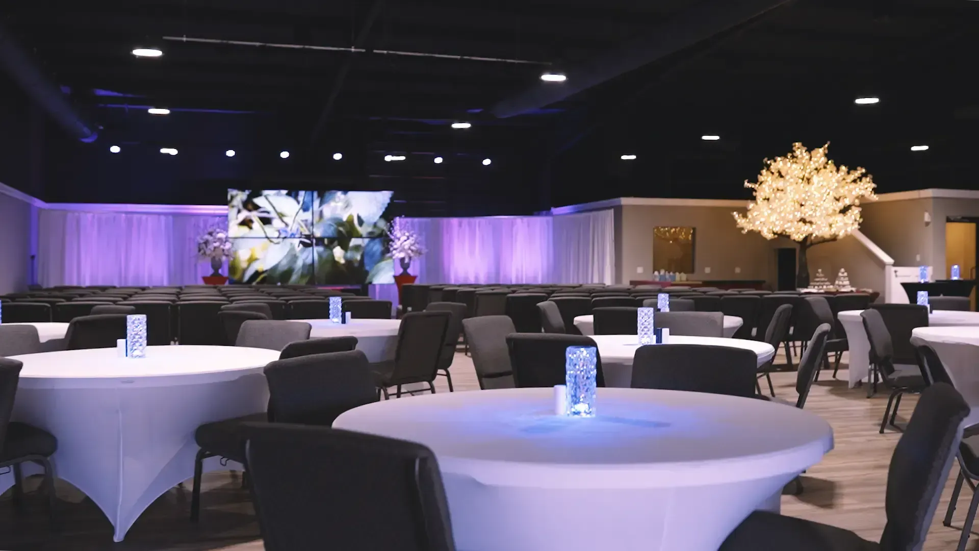 Banquet hall with round tables, black chairs, purple drapes, and two illuminated decorative trees