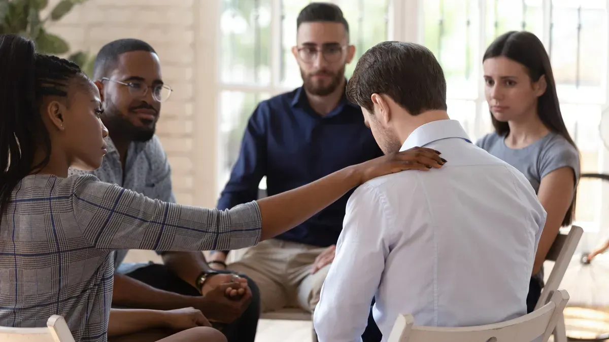 Support group seated in a circle, one person comforting another in a bright room.