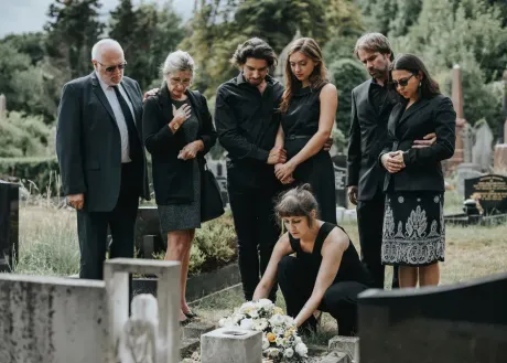 Mourners gather around a gravesite as a woman places flowers on a grave during a funeral.