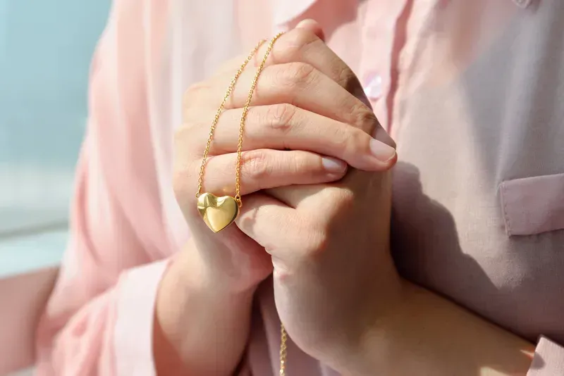 Hands holding a gold heart pendant on a chain against a soft pink shirt.