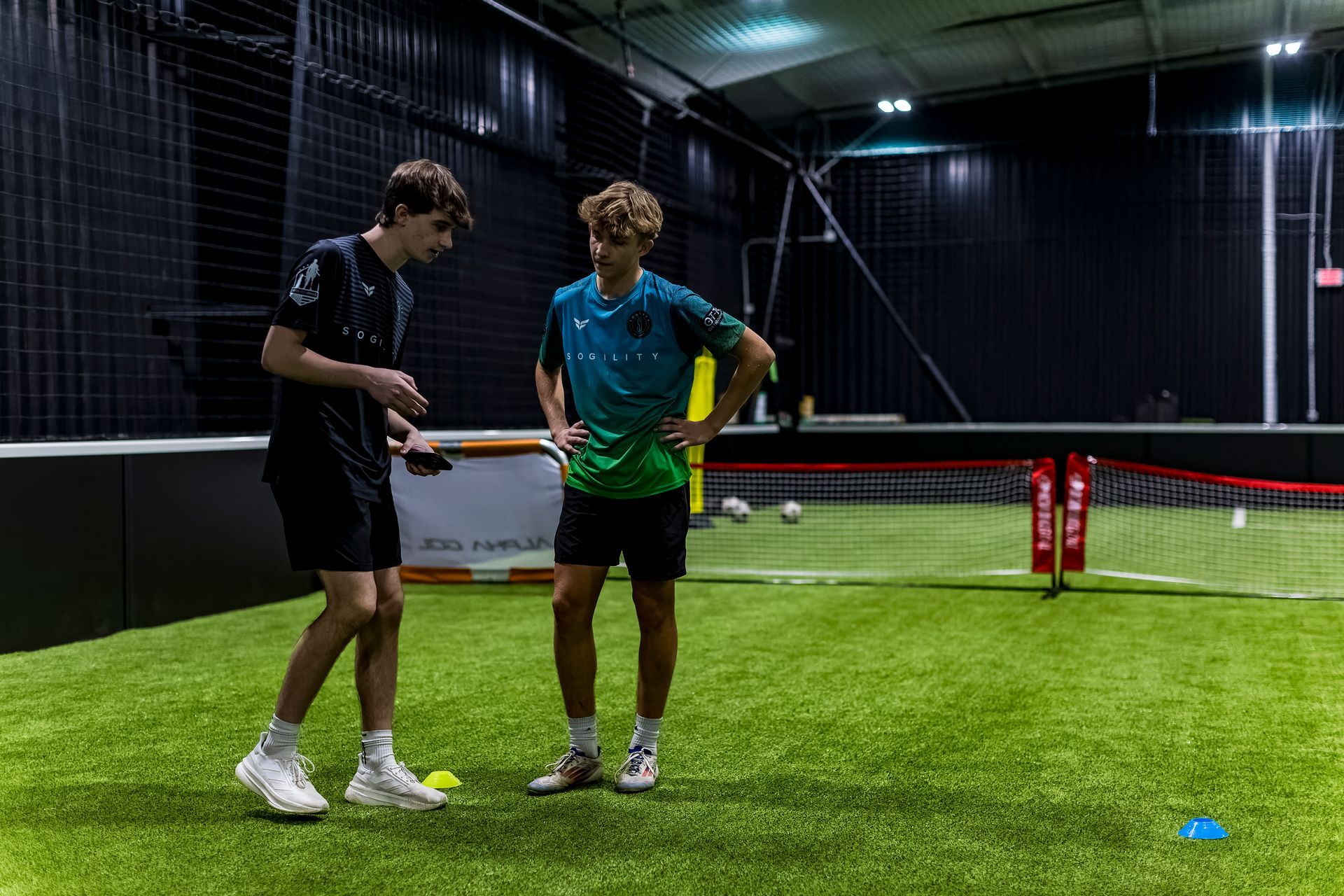 Two young men are standing on a soccer field talking to each other.