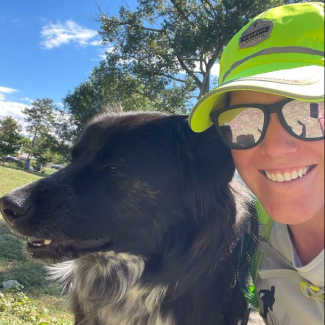 Woman smiling with a black and white dog in a sunny outdoor setting, both looking towards the left.