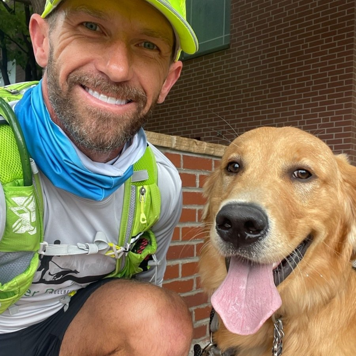 Man kneeling next to golden retriever, both smiling outdoors. Man wears running gear.