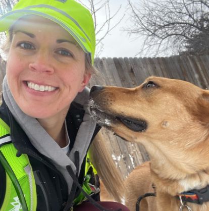 Woman smiles at camera with dog, snow on dog's snout. Outdoors, woman in neon vest, dog has brown fur.