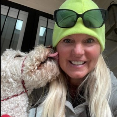 Woman smiling, dog licking her cheek. Woman wears a neon green beanie and sunglasses. Outdoors.