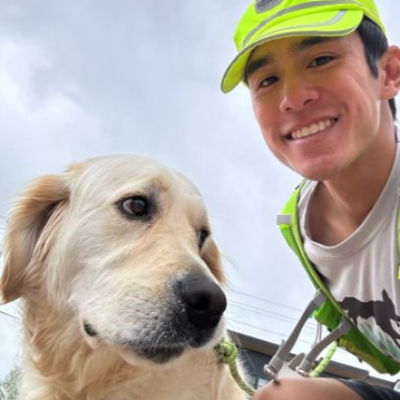 Man smiles, taking a selfie with a golden retriever dog outdoors, wearing a neon green vest and cap.