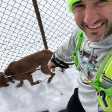 Man smiles for selfie with dog on leash in snow, wearing green vest and hat.