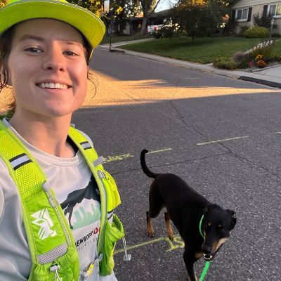 Emma in a reflective vest and hat smiles with a dog on a leash on a residential street.